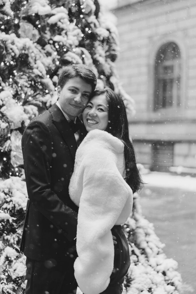Man in a tuxedo hugs his fiance wearing a silk dress and fur wrap in front of a snow covered Christmas tree in NYC